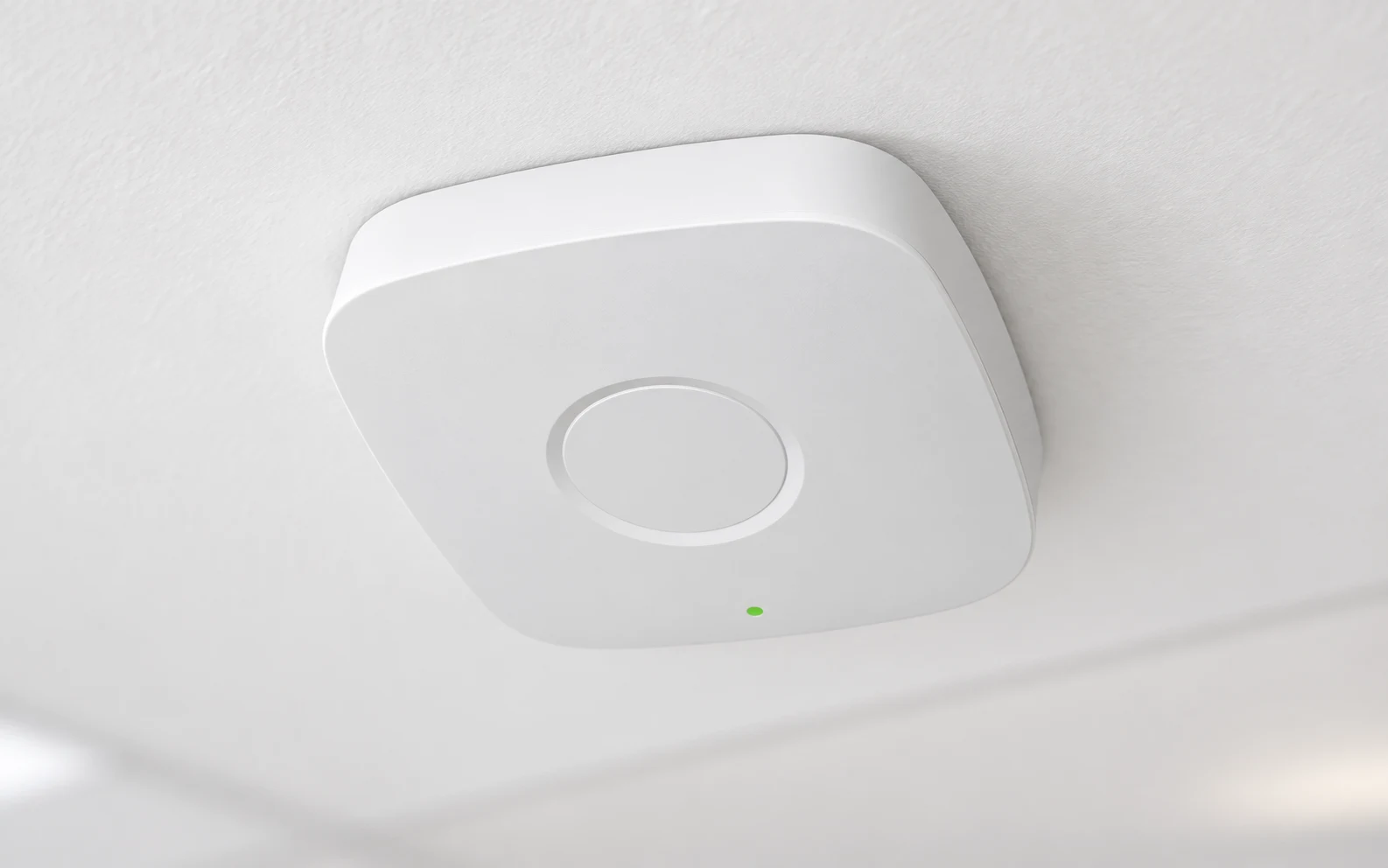 A white rounded-square ceiling access point seen from below, with a small green status light and an Ethernet port on its edge.